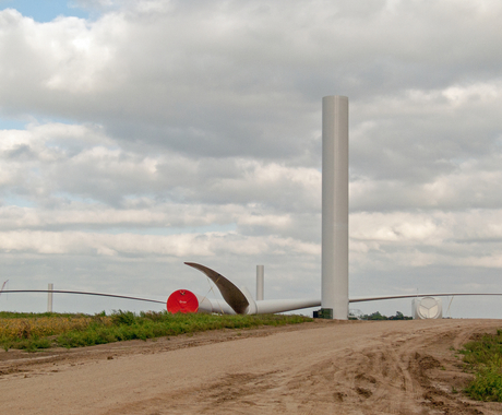 Photo of a wind turbine that is being constructed. The support structure, the base cylinder, is about halfway constructed. The blades are connected to the motor, but laying on the ground beside the base. A dirt road leads up to the site. In the background are two more partially constructed wind turbines, one with a crane beside it.