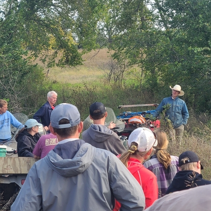 Men and women standing in a pasture. You can see the backs of people's heads and bodies with a man in a cowboy hat facing the crowd. 