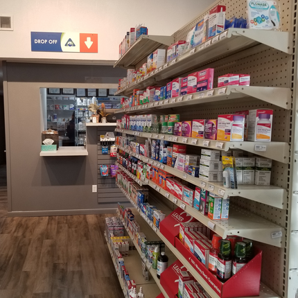Vista del interior de una farmacia que muestra una estantería repleta de diversos medicamentos sin receta, incluyendo medicamentos para la alergia, remedios para el resfriado y la gripe, y vitaminas. A la izquierda, hay un mostrador de entrega con un cartel que dice "DÉJATE" y símbolos direccionales. El mostrador tiene una pequeña estantería con una caja de pañuelos y algunos elementos decorativos, mientras que el área de trabajo de la farmacia, con estantes de medicamentos con receta, se ve a través de una ventana tras el mostrador. 