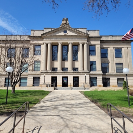 County government building with a sidewalk and green grass and flag pole in the foreground, and a blue sky. 