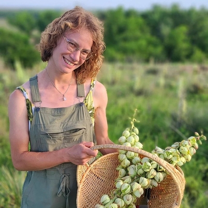 White woman with short, blond wavy hair, wearing glasses and green overalls holds a weaved basket with plants in it in a field of green plants behind her