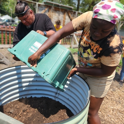 Una mujer negra vierte tierra de un contenedor verde en un contenedor grande y redondo de metal, mientras que un hombre de color con una camisa negra sostiene parte del contenedor verde trasero para ayudar