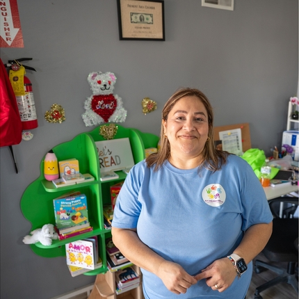 A woman wearing a light blue t-shirt stands in front of a bookshelf in a day care. 