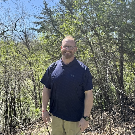 Man with thin hair on his head, dark glasses, and facial hair, wearing a blue polo shirt and khakis stands in a wooded area, smiling at the camera.