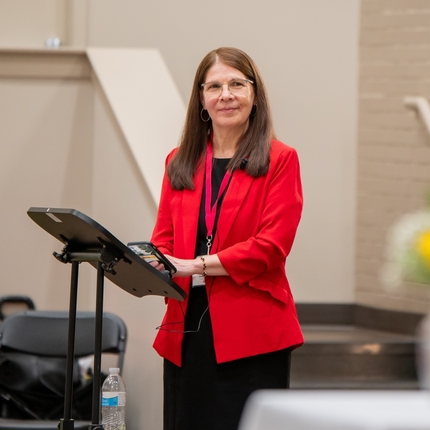 Woman with dark red hair wears a black shirt and black pants with a red suit jacket over it. She is standing at a podium with painted white brick walls behind her.