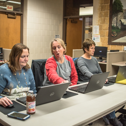 Three women sitting at a table, with three computers in front of them. The middle woman is looking at the left most computer, and pointing, while explaining something on the screen.