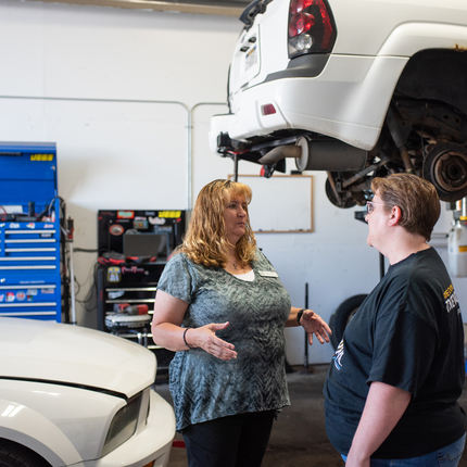 Two women standing in a car repair shop, in front of a car on the ground and an SUV elevated