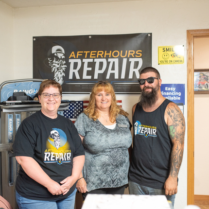 Three people - a woman on the right and man on the left wearing "Afterhours Repair" black shirts, and a woman in the middle with a short sleeved gray shirt, standing in an office in front of a sign that says "Afterhours Repair"