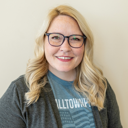 White woman with shoulder length blond hair wearing black rimmed glasses and a light blue shirt t-shirt with white wording and graphic design, and a dark gray cardingan over it.
