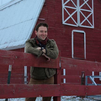 A smiling man in a green jacket and scarf leans on a red wooden fence in front of a large red barn with white trim. There is snow on the ground and the barn roof.