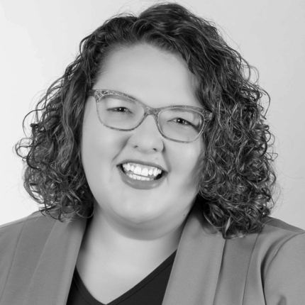 Woman smiling at the camera, with shoulder length curly brown hair, glasses, and a jacket over a dark shirt