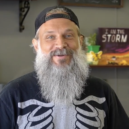 Man with full gray beard and backwards hat in T-shirt with rib cage on the front looking straight at camera