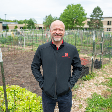Man with a bald head and facial hair, wearing a zip up collared black shirt with a red "N" on the left breast, smiles at the camera. He is standing in the middle of a garden with leafy greens and sprouts coming up, with fencing around each plot.