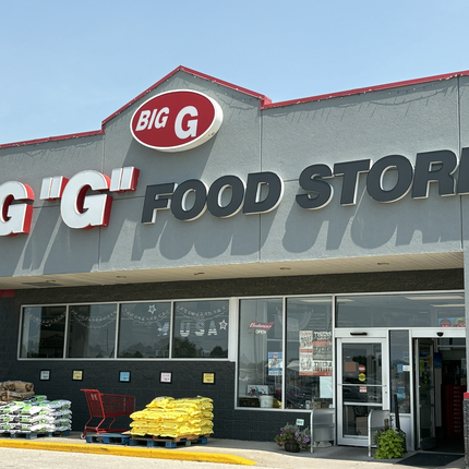 Front of a gray one-story building. Sign says "Big 'G' Food Store. Below the sign are large windows and two glass doors.
