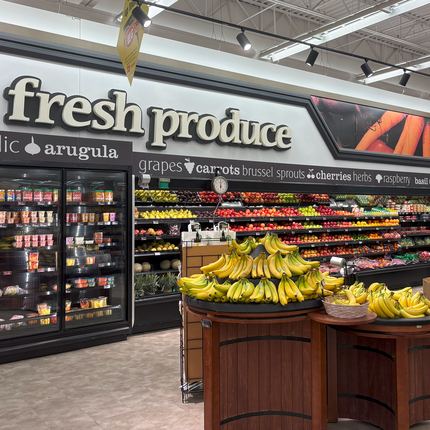 The words "fresh produce" are in white across the wall. Below them sit black coolers, stocked with apples, lemons, oranges, and a variety of vegetables. Toward the front of the photo we see bananas stacked on wooden risers. 