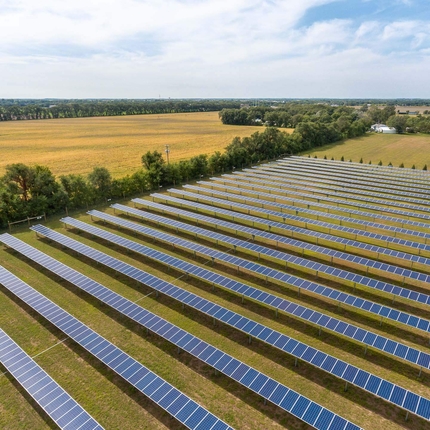 Vista aérea de hileras de paneles solares sobre una pradera verde, bordeada de árboles a ambos lados. Al fondo, un campo de cultivo y una granja.