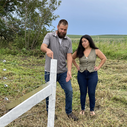 Un hombre y una mujer están de pie en un campo, en la esquina de un poste de cerca. El poste de cerca está hecho de una pala de turbina eólica reciclada y es de color blanco.