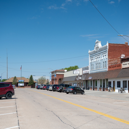 Photo looking down a small town Main Street. Diagonal parking is on each side of the street with a double yellow line running down the middle. On each side of the street are one and two story buildings with large storefront windows. At the end of the street is a sign that says "Blue Hill"