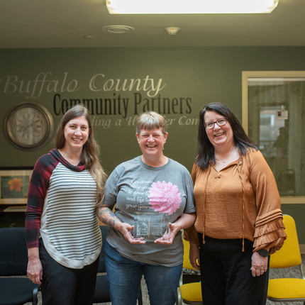 Three white females stand in a community room. The middle woman is holding an award plaque.