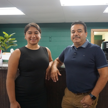 A young hispanic woman and an older hispanic man stand next to each other inside an office building with a green wall behind them and lights above, an American flag hangs behind the hispanic woman in the background