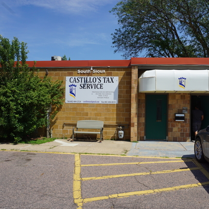 Brick building with an orange red trim around the top edge, a white sign with blue lettering on the front side of the building and a white overhang over two doors with a black car parked up front