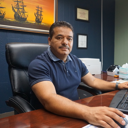 Hispanic male with black hair sits at a desk with a computer and keyboard in front of him, a painting of yellow ships with a yellow background hangs behind him on a wall