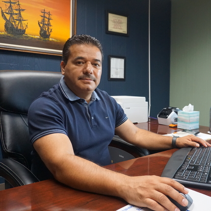 Hombre latino con camisa azul oscura esta sentado detrás de un escritorio con una computadora, y una pintura de barcos al fondo detrás de él