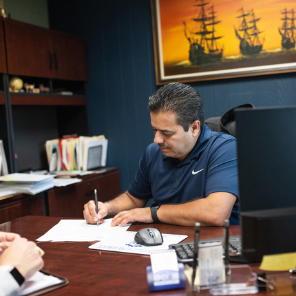 Un hombre hispano con cabello corto y oscuro, vestido con una camiseta polo Nike azul marino, detrás de un escritorio con una computadora, escribe algo en un papel blanco