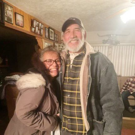 Retirement age Latino man and woman smiling warmly at the camera from the inside of their home, dressed for cold weather.