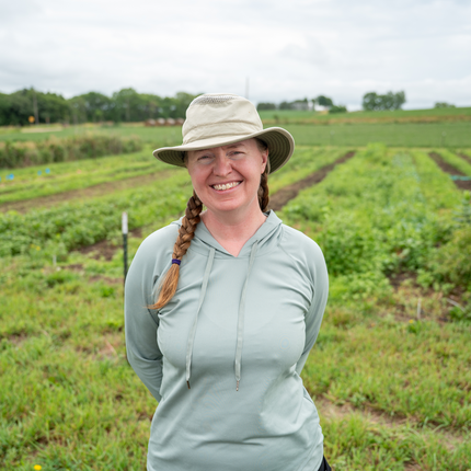 A woman wearing a mint green long sleeved shirt and a beige gardening hat stands in front of a lush green field.