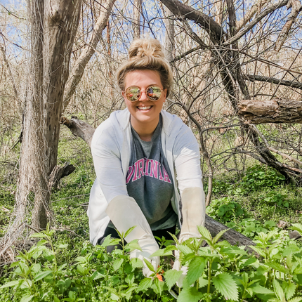 White woman with blond hair wearing sunglasses, white zip up hoodie sweater with a green shirt underneath is kneeled down in greenery