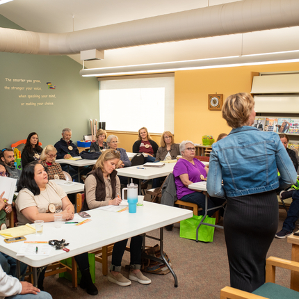 Group of more than 10 men and women sit at long tables facing towards a white woman with short blond hair wearing a jean jacket and black dress presenting to the group in a medium size room with green and yellow walls