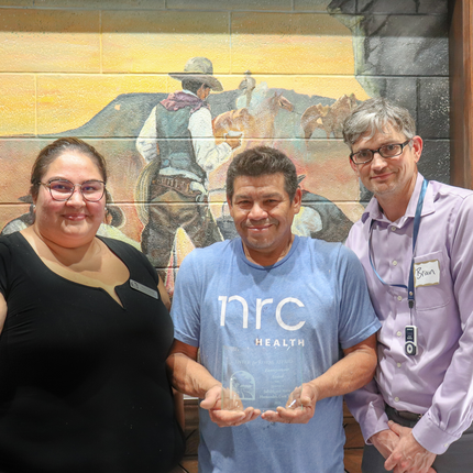Un grupo de tres personas posa en un interior frente a un colorido mural con una escena de vaqueros. El hombre del centro lleva una camiseta azul claro de "NRC Health" y sostiene un premio de cristal transparente. La mujer de la izquierda lleva gafas y una camiseta negra, mientras que el hombre de la derecha lleva gafas, una camiseta morada clara, una etiqueta con el nombre "Brian" y un cordón.