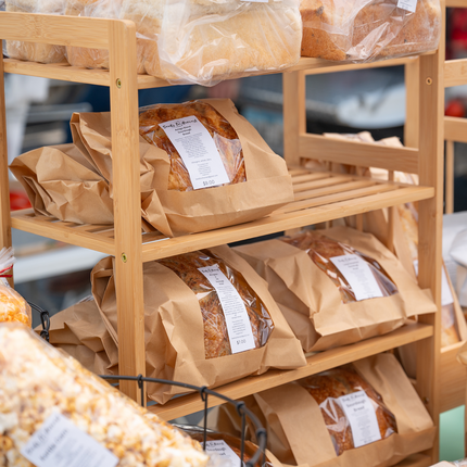 A wooden rack of an assortment of breads, wrapped in clear cellophane and brown parchment. This photo appears to be from a table at a farmers market.