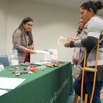 Latina woman with long dark hair and glasses stand behind a table with a green table cloth handing a paper to two women standing in front of her, both hispanic women, one holding a brown crutch under her arm 