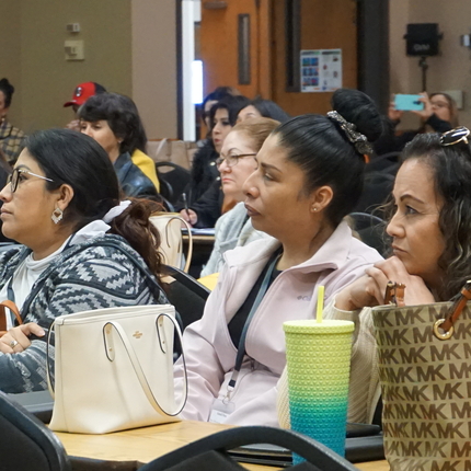 3 mujeres latinas sentadas en una mesa con sus bolsos frente a ellas, rodeadas por una sala llena de gente en una conferencia