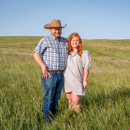 A man and woman stand together in a grassy field under a clear blue sky. The man wears a cowboy hat, plaid shirt, jeans, and boots, while the woman wears a light-colored dress with turquoise jewelry and cowboy boots. They are smiling and standing close to each other.