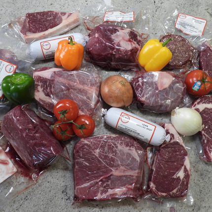 A few cuts of packaged beef arranged in a circle on a table. Cuts are in clear packaging with a couple of white packages of ground beef. Arranged throughout are fresh peppers, tomatoes, and onions.