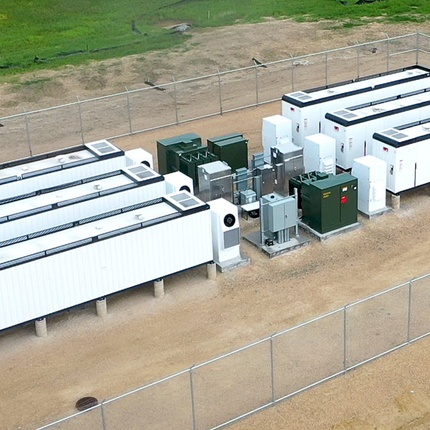 Battery storage project- six small white rectangle buildings (about the size of tractor trailers) lined up three on each side with green and gray electric junction boxes in between. All are in a chain linked fence area with brown dirt underneath.