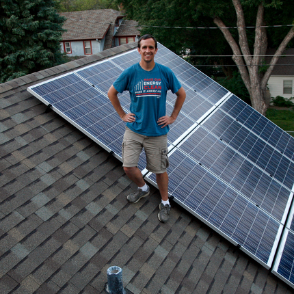 Un hombre blanco de pelo oscuro se encuentra de pie sobre un tejado de tejas grises, frente a unos paneles solares recién instalados. Sonríe con las manos en las caderas, lleva una camiseta azul claro que dice "Hagamos que nuestra energía sea limpia, que sea estadounidense" y pantalones cortos color caqui.