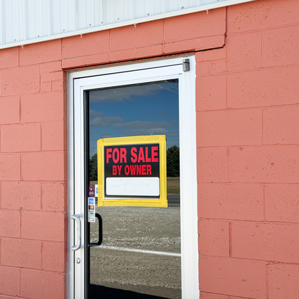 Salmon-colored brick building with a clear business door in middle. A "for sale by owner" sign in the top center of the door.