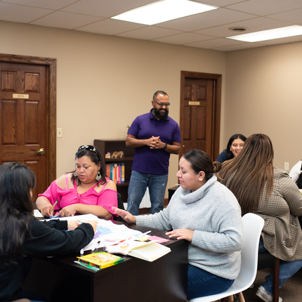 Latina women working together at tables of three, with a man in a purple shirt standing, smiling, and assisting