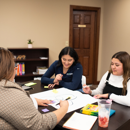 Three Hispanic women sit at a dark table where they are drawing a diagram on a large white paper with different colored markers, there is a brown door behind them.