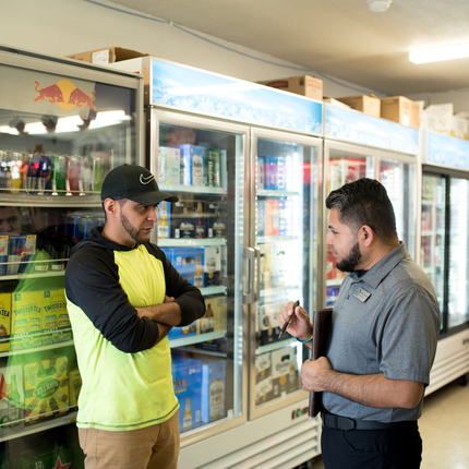 two men talking to each other, standing in front of coolers