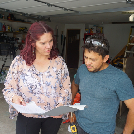Hispanic woman with red hair wearing a light pink flowered shirt and black pants holds white papers while showing them to a hispanic male wearing a navy shirt and protective glasses on his head outside of a garage