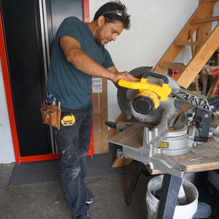 Hispanic man wearing a navy short sleeve shirt, black pants and a tool belt around his waist uses a big cutting tool to cut a wood flooring panel inside a garage