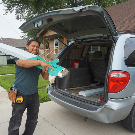 Hispanic man wearing a navy shirt and black jeans carries box of white box of wood flooring panels out of the back of a light blue van outside a neighborhood