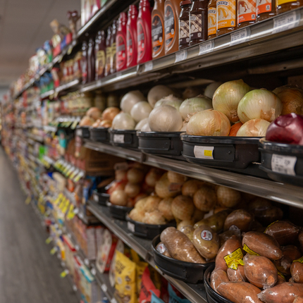 Looking down one side of a grocery store aisle. In the foreground are four types of onions on one shelf, and different kinds of potatoes underneath. Above the onions are ice cream syrups. 
