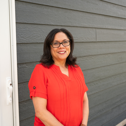 Woman in a short sleeved, red/orange blouse standing in front of a white door and gray siding. She is smiling warmly and looking into the camera.