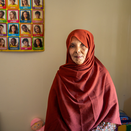 A dark-skinned older woman wearing a maroon hijab, looking directly at the camera. She is standing in front of a beige wall with a white door to her left (right side of the photo). On the left side is a poster hanging up that says "Emotions" and has photos of children and words that state emotions in each photo.